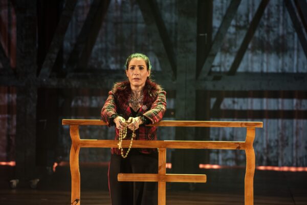 A imagem mostra uma cena de teatro em que uma mulher está em pé, apoiada em uma estrutura de madeira que parece ser um tipo de balcão ou suporte. Ela está usando uma roupa predominantemente vermelha, com um casaco xadrez e detalhes em pele ao redor da gola. A expressão dela é séria e introspectiva, como se estivesse absorvida em pensamentos profundos ou num momento de tensão. Em suas mãos, ela segura um rosário ou contas de oração, o que pode sugerir que a personagem está em um momento de reflexão ou oração. Ao fundo, a iluminação é suave, destacando a personagem, enquanto a estrutura metálica e sombria ao fundo sugere um cenário de ambiente fechado, possivelmente uma prisão ou um local com uma atmosfera opressiva.